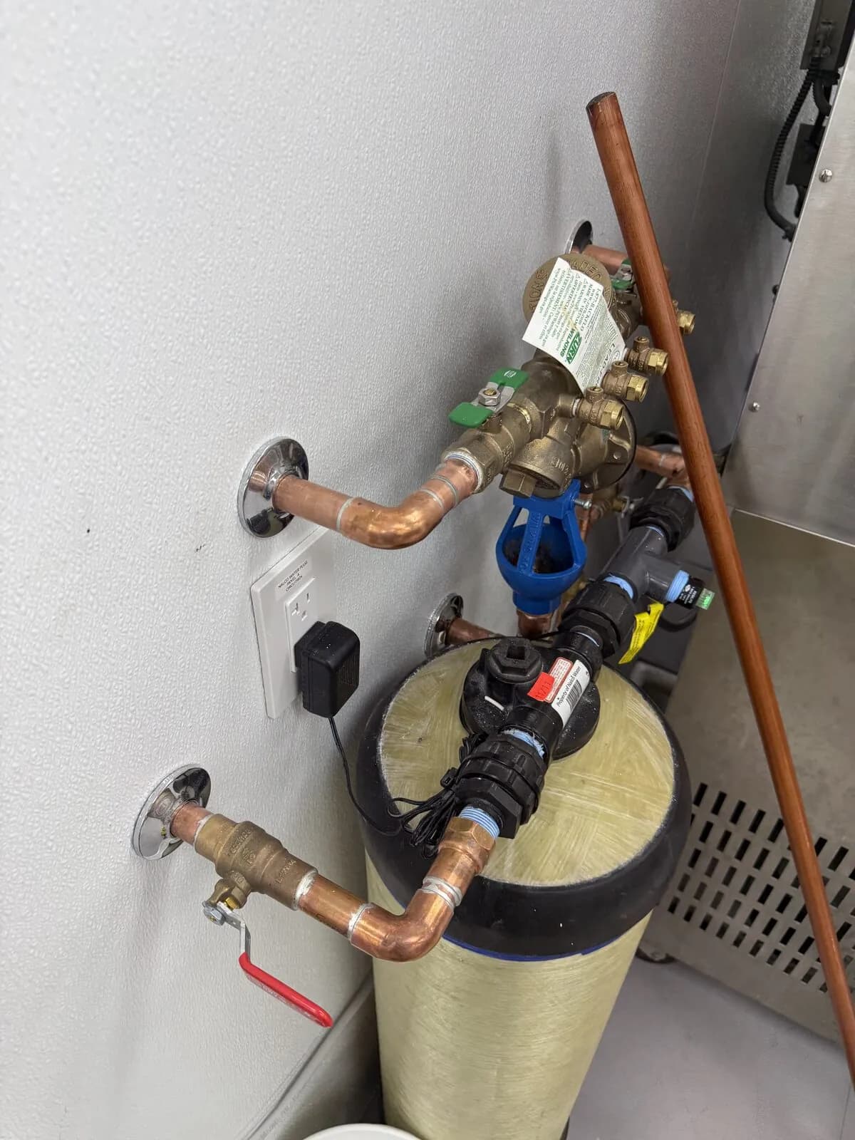 Commercial water filtration tank with copper piping, shutoffs, and backflow assembly on a white wall
