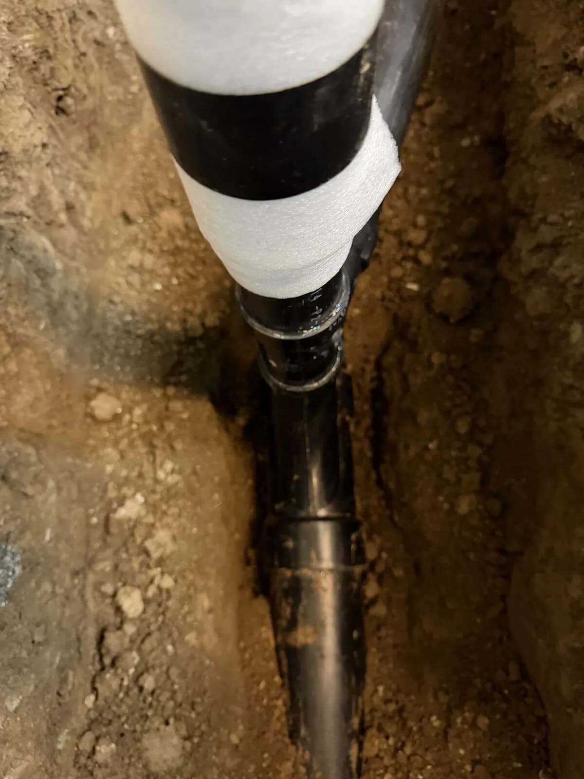 Insulated black underground drain pipe wrapped and taped inside a narrow earthen trench
