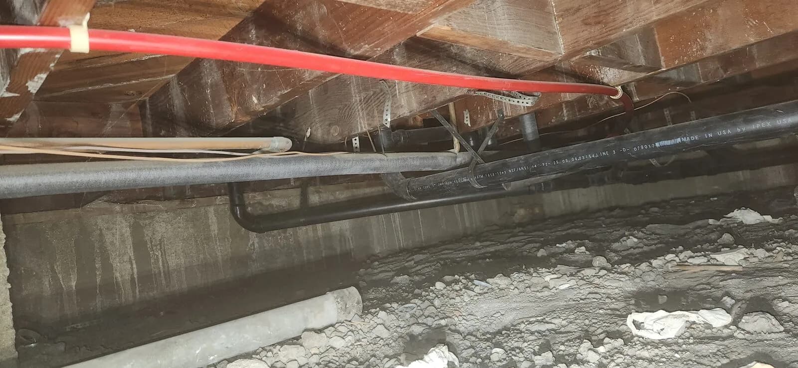 Red PEX water lines and black ABS drains routed under floor joists in a crawl space