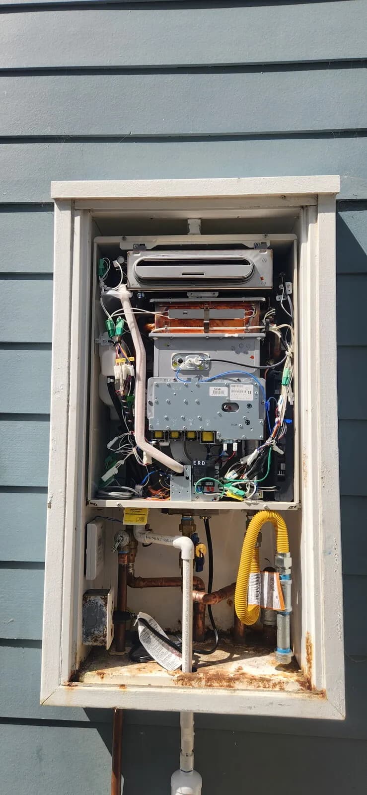 Outdoor tankless water heater with cover removed showing wiring, heat exchanger, and plumbing connections